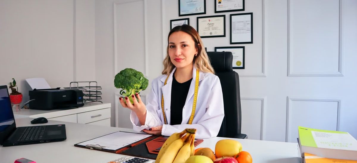 Female nutritionist in office holding broccoli, surrounded by fruits, promoting healthy lifestyle.