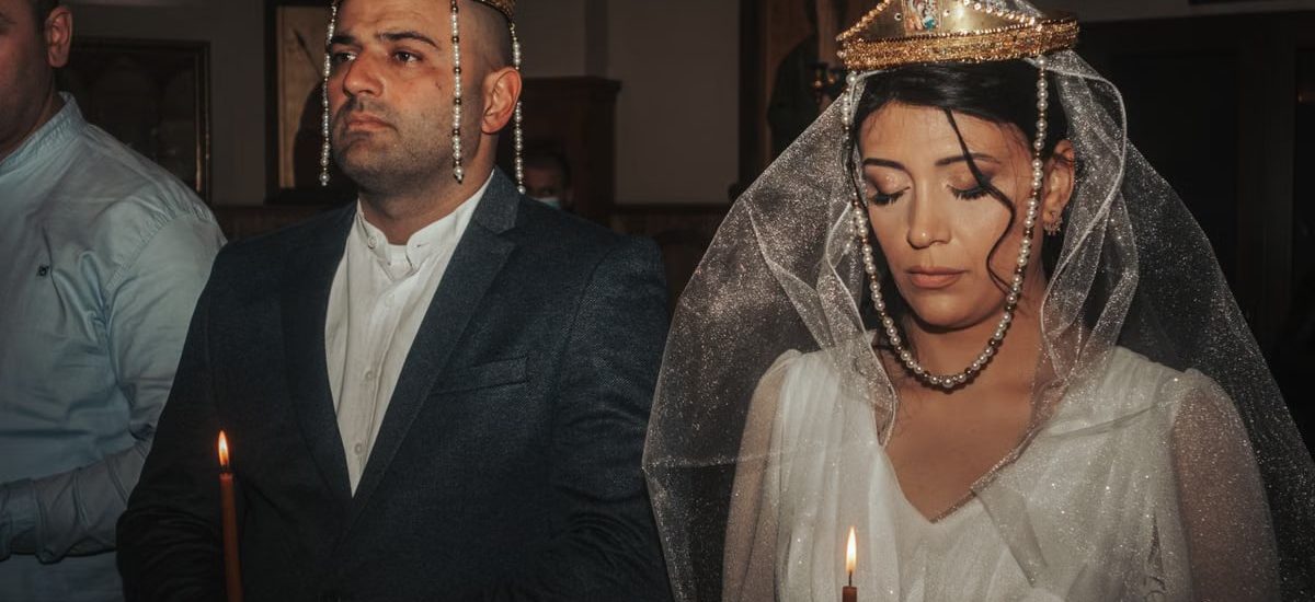 Bride and groom in traditional wedding attire with crowns, holding candles in solemn ceremony.
