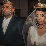Bride and groom in traditional wedding attire with crowns, holding candles in solemn ceremony.