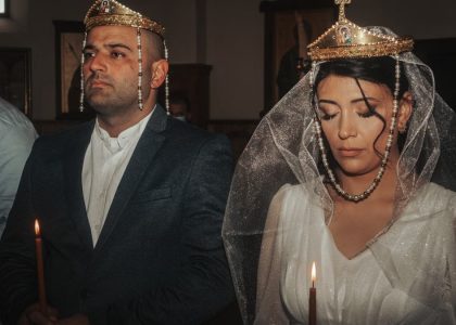 Bride and groom in traditional wedding attire with crowns, holding candles in solemn ceremony.